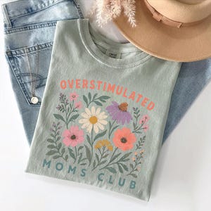 a green t-shirt with a floral design, a pair of jeans, and a beige hat, all laid out on a white surface.