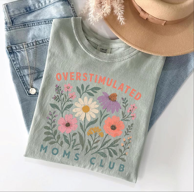 a green t-shirt with a floral design, a pair of jeans, and a beige hat, all laid out on a white surface.