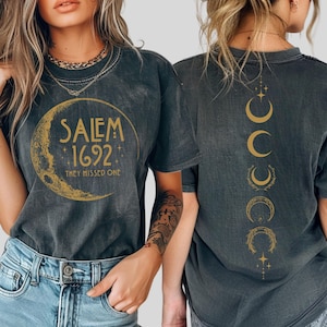 May include: Dark gray t-shirt with a gold crescent moon graphic that reads "Salem 1692 They Missed One." The back of the shirt features a vertical design of gold crescent moons and stars. The shirt is worn with blue jeans.
