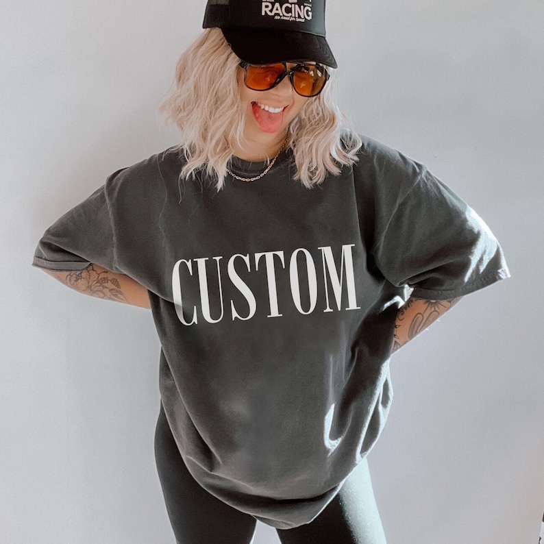 A smiling woman wearing a grey t-shirt with the word "CUSTOM" printed on it, along with a black cap and sunglasses.