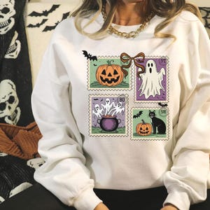A person wearing a white sweatshirt with a Halloween-themed design featuring a jack-o-lantern, a ghost, a witch's cauldron, and bats.