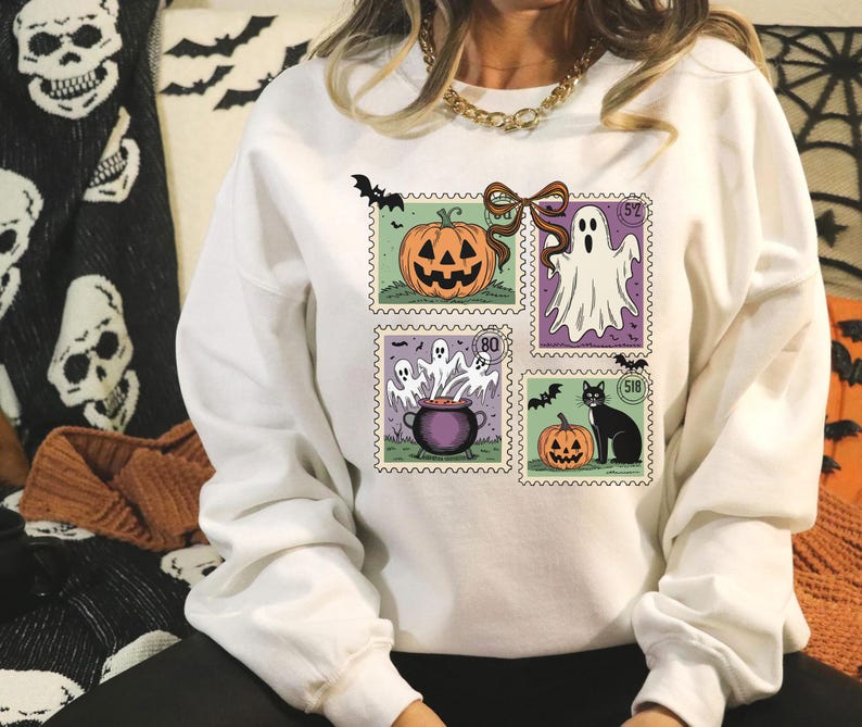 A person wearing a white sweatshirt with a Halloween-themed design featuring a jack-o-lantern, a ghost, a witch's cauldron, and bats.