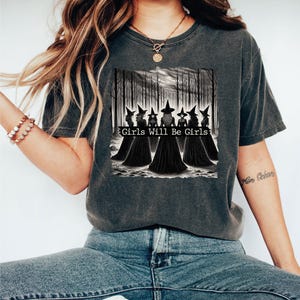 May include: Dark grey t-shirt featuring a graphic of six witches in black robes and pointed hats, standing in a forest. The text "Girls Will Be Girls" is printed below the image. The shirt is made of a soft material.