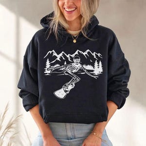 A woman wearing a black hoodie with a graphic design of a skeleton riding a snowboard against a mountain landscape.
