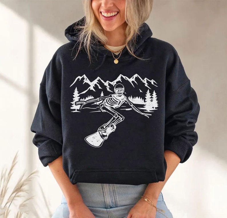 A woman wearing a black hoodie with a graphic design of a skeleton riding a snowboard against a mountain landscape.