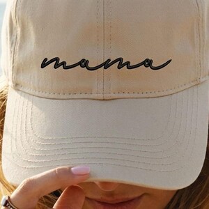 A woman wearing a beige baseball cap with the word "mama" printed on it, looking over her hat with her hand.