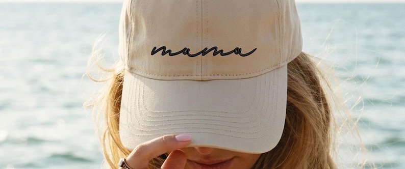 A woman wearing a beige baseball cap with the word "mama" printed on it, looking over her hat with her hand.