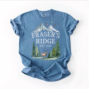 A blue t-shirt with a graphic design depicting a mountain landscape, featuring a deer and pine trees. The text on the t-shirt reads "Fraser's Ridge" and "EST. 1767".