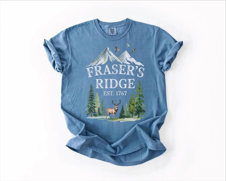 A blue t-shirt with a graphic design depicting a mountain landscape, featuring a deer and pine trees. The text on the t-shirt reads "Fraser's Ridge" and "EST. 1767".
