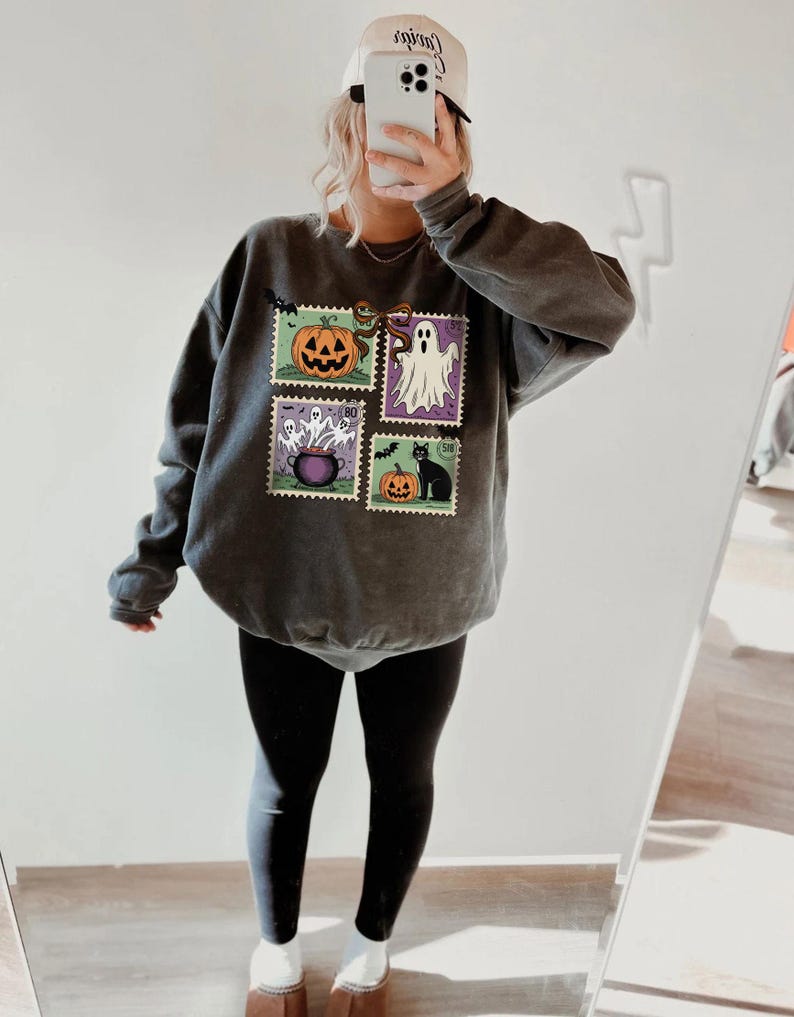 A person is taking a selfie in front of a mirror, wearing a gray sweatshirt with Halloween-themed designs and a white baseball cap.