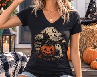 Può includere: Una t-shirt nera con scollo a V caratterizzata da una stampa grafica raffigurante una zucca, un fantasma con cappello da strega che tiene una tazza "BOO", una casa stregata, pipistrelli e un cartello con scritto "SPOOKY VIBES" su una luna gialla.