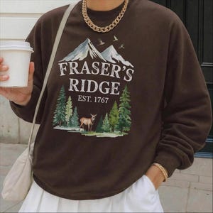 A person wearing a brown sweatshirt with a graphic design of mountains, pine trees, and a deer, holding a white coffee cup.