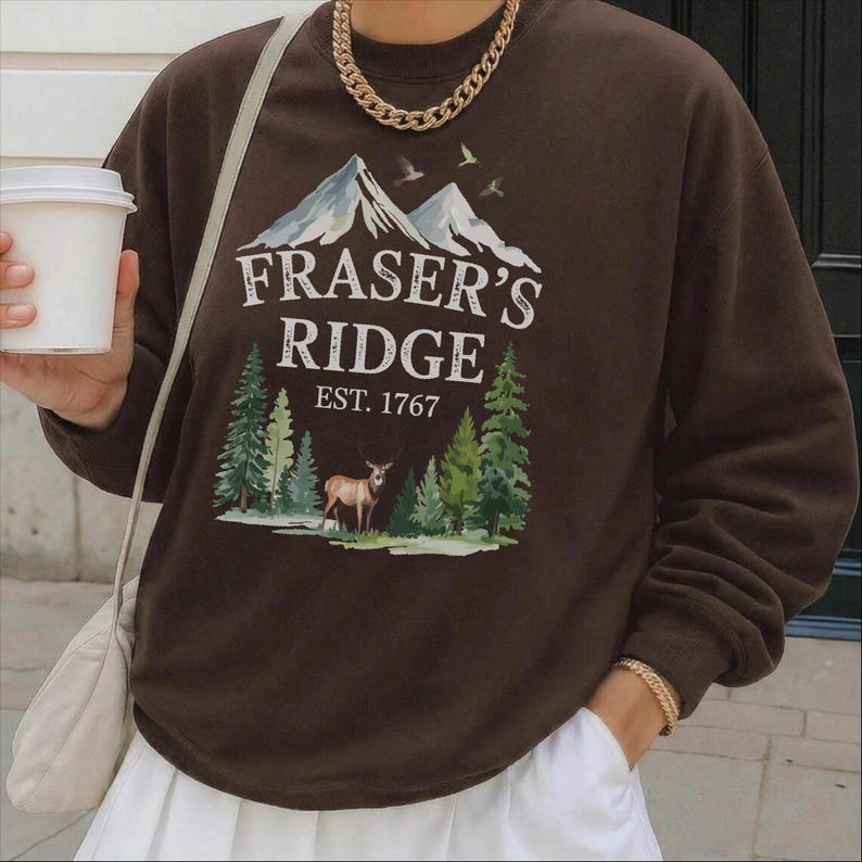 A person wearing a brown sweatshirt with a graphic design of mountains, pine trees, and a deer, holding a white coffee cup.