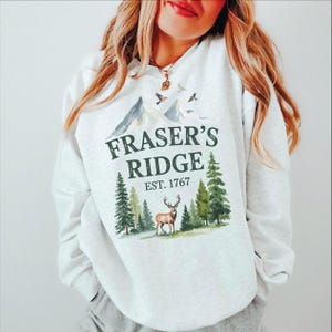 A woman wearing a white sweatshirt with a graphic design featuring a deer and mountains, along with the text "Fraser's Ridge Est. 1767".
