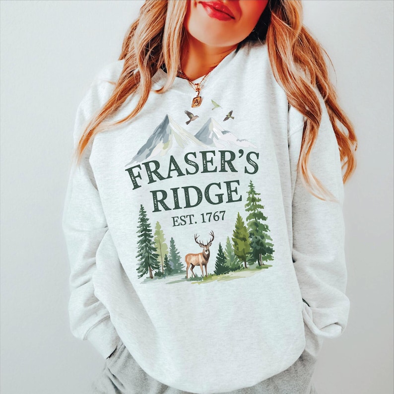 A woman wearing a white sweatshirt with a graphic design featuring a deer and mountains, along with the text "Fraser's Ridge Est. 1767".