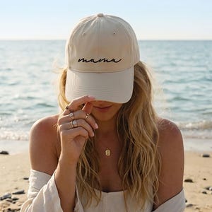A woman wearing a beige cap with the word "mama" on it, standing on a beach with the ocean in the background.