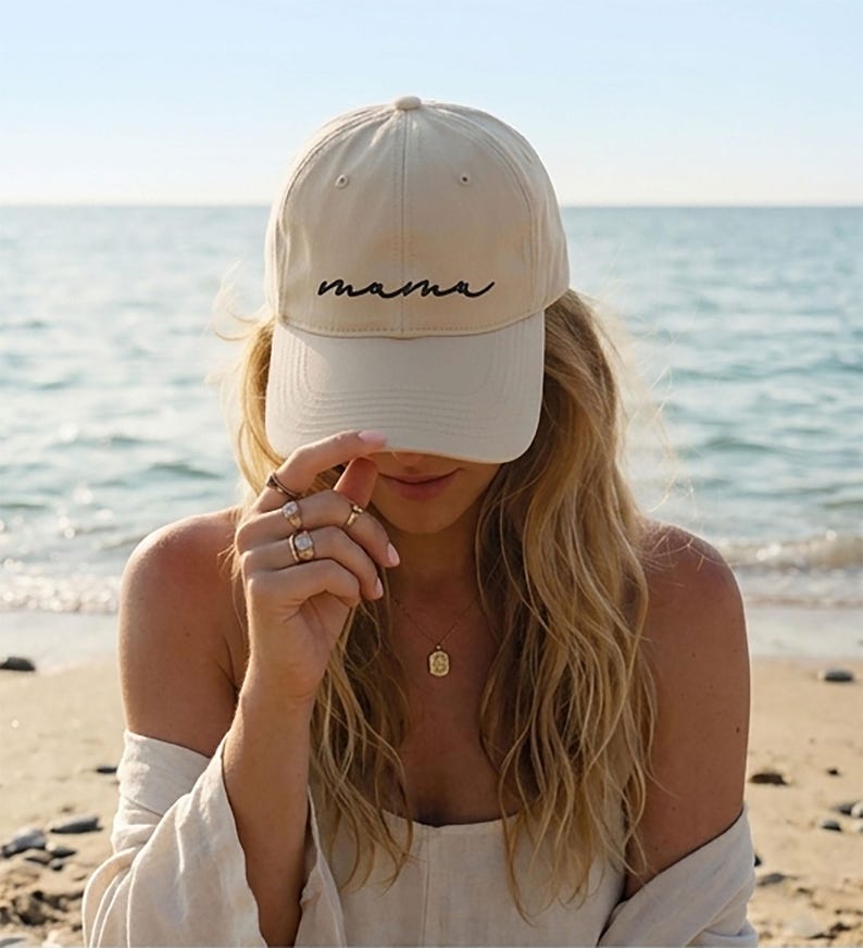 A woman wearing a beige cap with the word "mama" on it, standing on a beach with the ocean in the background.