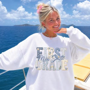 May include: A white crewneck sweatshirt featuring the text "FIRST GRADE" in large, blocky letters with a blue and cream floral and gingham pattern. The words "Miss Smith" are written in a cursive blue script across the center of the design.