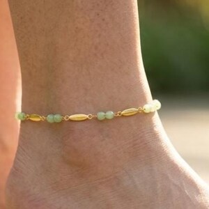 May include: An anklet featuring a delicate gold chain interspersed with small, smooth, light green beads and elongated gold-toned metal spacers. The jewelry piece wraps around the ankle, showcasing a repeating pattern of beads and metal links.
