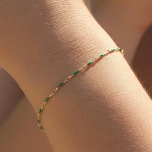 May include: A delicate gold bracelet with alternating green and gold links is shown on a person's arm. The bracelet is thin and elegant, with a simple design. The background is blurred, suggesting an outdoor setting.