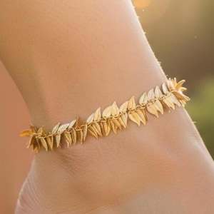 May include: A gold-colored anklet featuring a leaf design. The anklet is made of small, connected leaf-shaped elements. The jewelry is worn on an ankle, with a blurred background of green and sunlight.