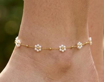 May include: A delicate gold anklet adorned with small, white flower-shaped beads and tiny gold beads. The anklet is worn on a foot, with a blurred green background suggesting an outdoor setting. A simple and elegant piece of jewelry.