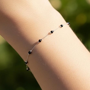 May include: Delicate bracelet featuring small, black beads strung along a thin, silver chain. The bracelet is displayed on a person's wrist, with a blurred green background suggesting an outdoor setting. A simple and elegant piece of jewelry.