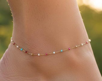 May include: A delicate gold anklet adorned with small, colorful beads. The anklet features red, turquoise, and white beads, strung along a fine gold chain. The jewelry is displayed on a person's ankle, with a blurred green background.