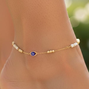 May include: A gold-colored anklet with small white beads and a blue and white evil eye charm. The anklet is worn on a foot, with a blurred green background.