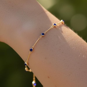 May include: A delicate gold bracelet with small, round, deep blue gemstones evenly spaced along the chain. The bracelet is shown on a person's wrist, with a blurred green background.