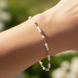 May include: Delicate gold-toned bracelet with small, white, round beads. The bracelet is strung on a fine chain and is worn on a wrist. The background is blurred with green and white tones.