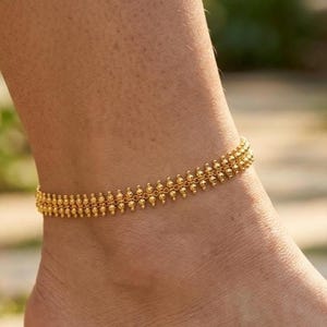 May include: A gold-colored anklet with a beaded design. The anklet is worn on an ankle and has a delicate, intricate pattern. The jewelry is likely made of metal and is a fashion accessory.