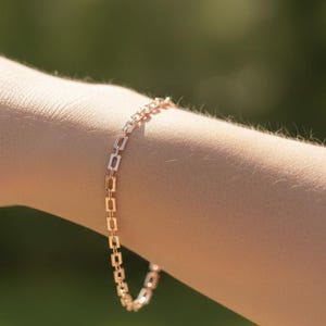 May include: Rose gold bracelet featuring a delicate rectangular link design. The bracelet is shown on a person's arm, with a blurred green background. The jewelry piece is a simple yet elegant accessory.