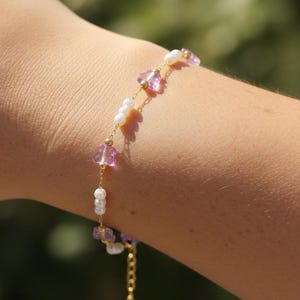 May include: Delicate gold-toned bracelet featuring alternating pink and white beads. The bracelet has a delicate chain and a small dangling charm. The bracelet is worn on a person's arm.