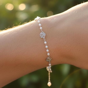 May include: A delicate bracelet featuring small white pearls and silver-toned floral accents. The bracelet is adorned with three decorative floral charms and a dangling pearl pendant. The jewelry is displayed on a person's wrist.