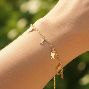 May include: A delicate gold-colored bracelet featuring small fish charms. The bracelet is worn on a wrist, with a blurred green background. The fish charms are evenly spaced along the chain, adding a whimsical touch to the jewelry.