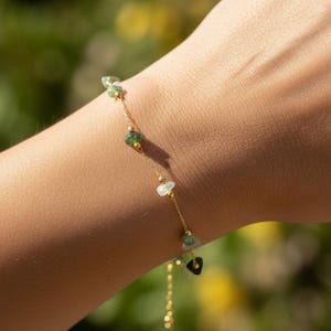 May include: A delicate gold bracelet adorned with small, irregularly shaped green and white gemstones. The bracelet is strung on a fine gold chain and is displayed on a person's arm against a blurred green background.