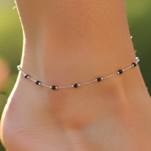 May include: A delicate silver anklet featuring small, round black beads evenly spaced along a fine chain. The anklet is shown on an ankle, with a blurred green background.