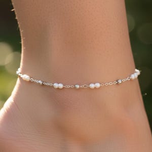 May include: A delicate anklet featuring small, round white pearls and silver-colored beads strung on a fine chain. The anklet is worn on an ankle, with a blurred green background.