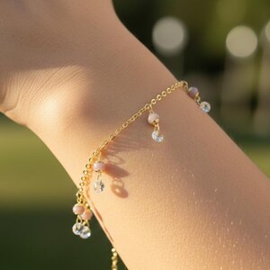 May include: A delicate gold bracelet with dangling charms. The bracelet features small, round pink beads and clear, sparkling crystals. The chain is thin and elegant, perfect for everyday wear. The bracelet is shown on a person's wrist.