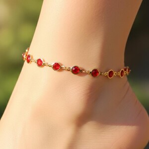 May include: A gold-colored anklet with red gemstones. The anklet is made of small, round red stones set in a gold chain. The anklet is worn on the ankle. The background is blurred, with green and brown tones.