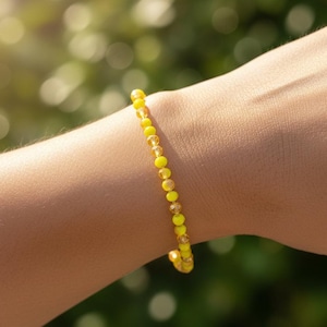 May include: A yellow beaded bracelet with small, round beads. The bracelet is worn on a person's arm, with a blurred green background. The beads are a mix of yellow and clear, creating a delicate and bright accessory.