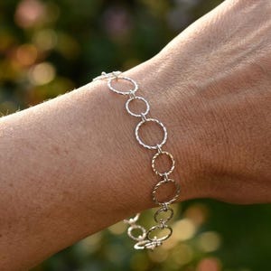 May include: Silver bracelet featuring a series of interconnected, textured circles. The bracelet is worn on a wrist, with a blurred green and gold background. The circles have a mix of smooth and twisted textures, creating a delicate and elegant design.