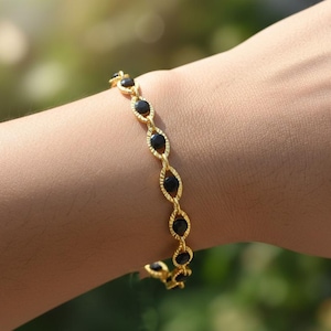 May include: A gold-toned bracelet featuring a series of interconnected, textured oval links, each set with a small, round, polished black stone. The chain design creates a repeating pattern of metal frames surrounding the dark circular inlays.