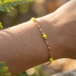 May include: A delicate gold chain bracelet featuring small, vibrant neon yellow beads spaced evenly along the twisted metal links. The bracelet is worn around the wrist, showcasing a simple design with alternating gold and bright yellow accents.