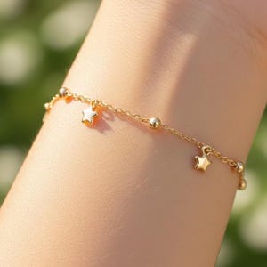 May include: A delicate gold bracelet featuring small star charms and tiny ball accents. The bracelet is shown on a wrist, with a soft, blurred green background. The bracelet's chain is thin and the stars are evenly spaced.