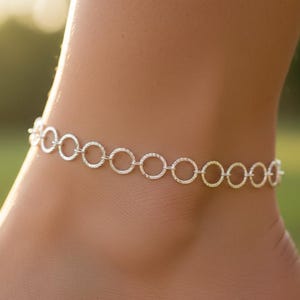 May include: A delicate silver anklet featuring a series of interconnected circles. The anklet is worn on the ankle, with a blurred background of green and yellow hues. The circles have a textured surface.