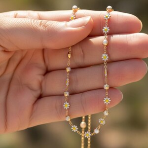 May include: A delicate gold necklace featuring small white daisy flower pendants with yellow centers and pearl accents. The necklace is held in a hand, showcasing its intricate design and craftsmanship. The chain is thin and elegant.