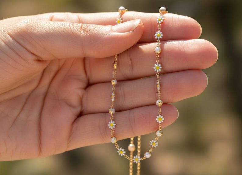 May include: A delicate gold necklace featuring small white daisy flower pendants with yellow centers and pearl accents. The necklace is held in a hand, showcasing its intricate design and craftsmanship. The chain is thin and elegant.