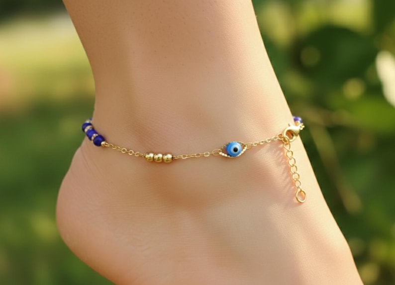 May include: A gold-tone chain anklet featuring a series of small blue beads, three gold-tone spherical beads, and a single blue and white evil eye charm. The jewelry piece includes a lobster claw clasp and an adjustable extender chain for sizing.